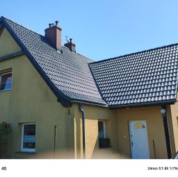 Firma ogólno-budowlana PawBud - Dom jednorodzinny z ciemną dachówką w słoneczny dzień. Widoczne kominy, okna z firankami i elewacja w odcieniach beżu. Ujęcie z dołu, podkreślające geometrię dachu.