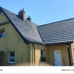 Dom jednorodzinny z ciemną dachówką w słoneczny dzień. Widoczne kominy, okna z firankami i elewacja w odcieniach beżu. Ujęcie z dołu, podkreślające geometrię dachu.