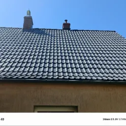 Ciemnoszara dachówka ceramiczna na skośnym dachu domu jednorodzinnego, widoczne dwa kominy z elementami wentylacyjnymi, błękitne niebo w tle.