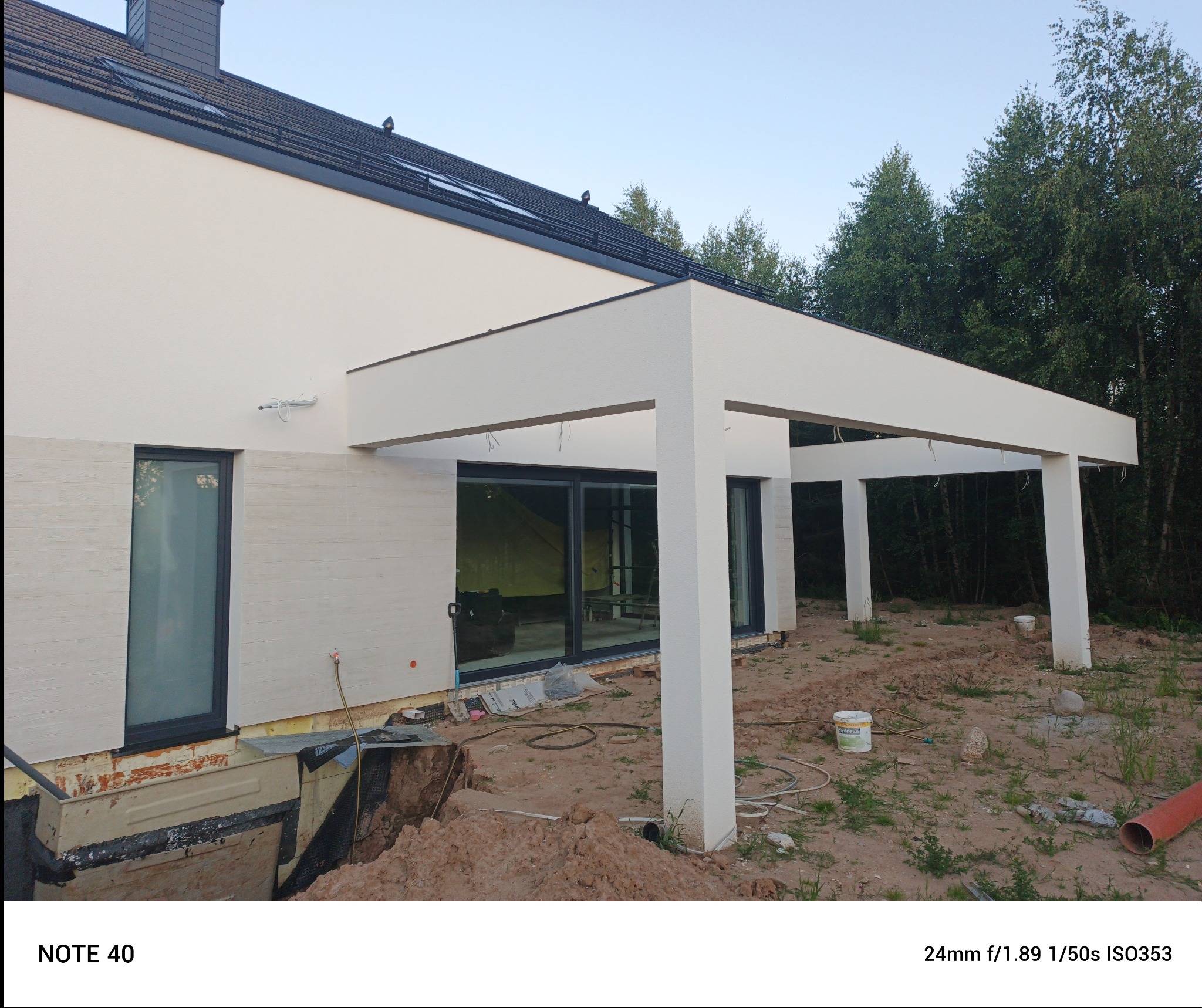 Elewacja domu z białym, nowoczesnym zadaszeniem tarasu. Widoczne prace ziemne i fragment dachu. Ujęcie z dołu ukazuje detale wykończenia.