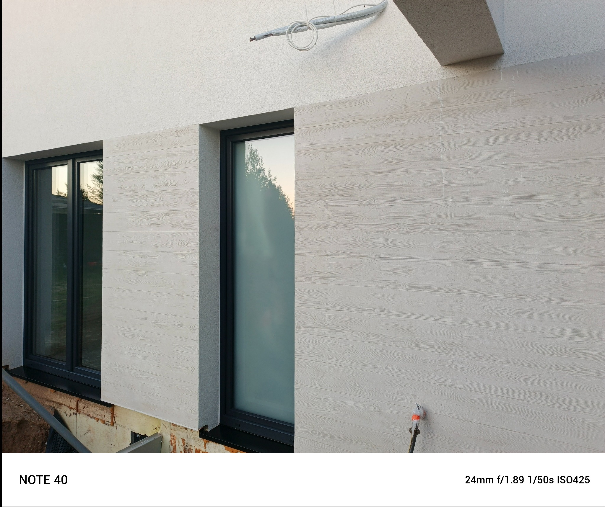 Elewacja budynku z oknami, wykończona panelami imitującymi drewno w jasnym odcieniu. Widoczne detale wykończenia i instalacja na ścianie.