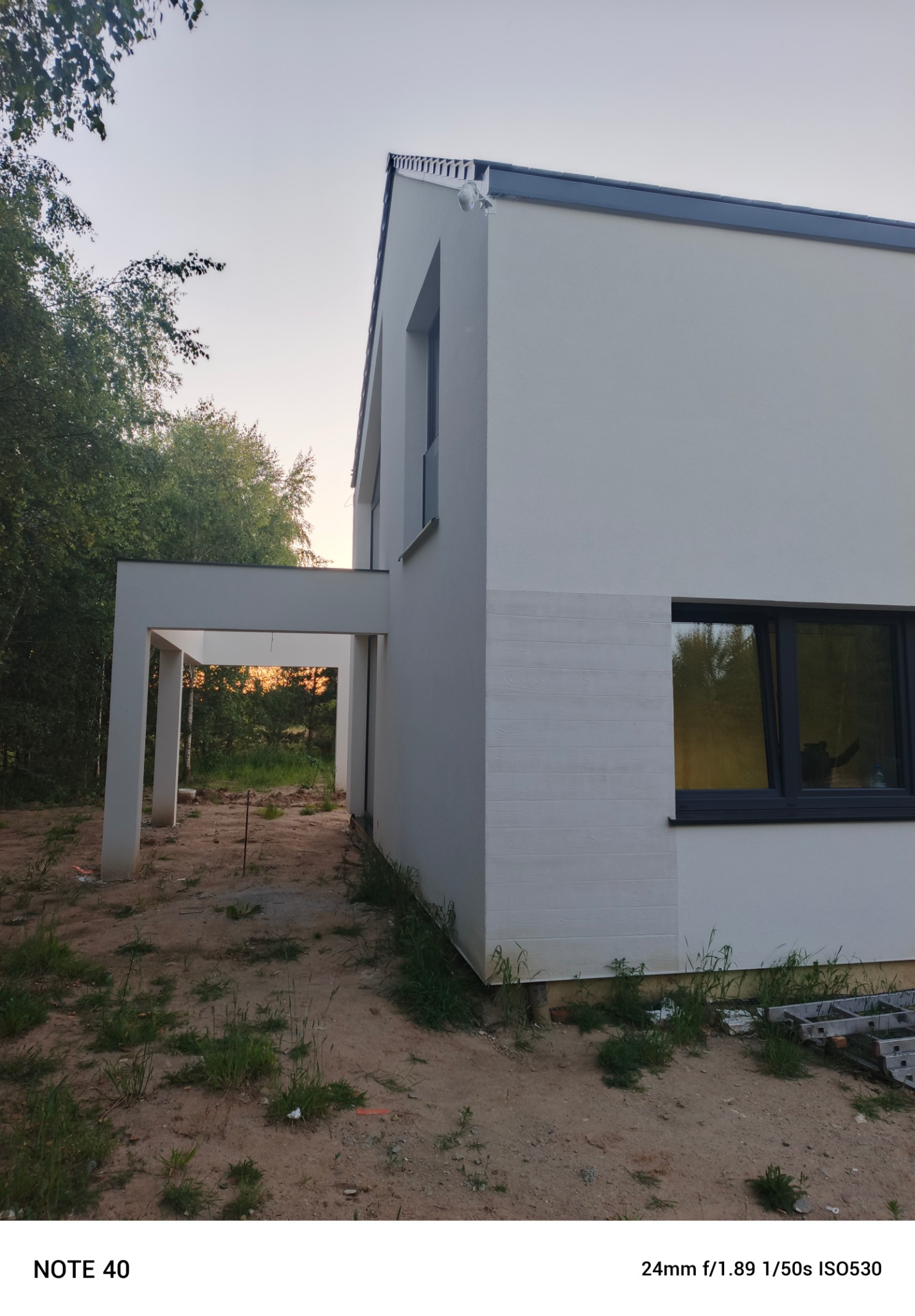 Nowoczesny, minimalistyczny dom z jasną elewacją i ciemnymi oknami, widziany z boku na tle zieleni. Detal elewacji imitujący drewno.