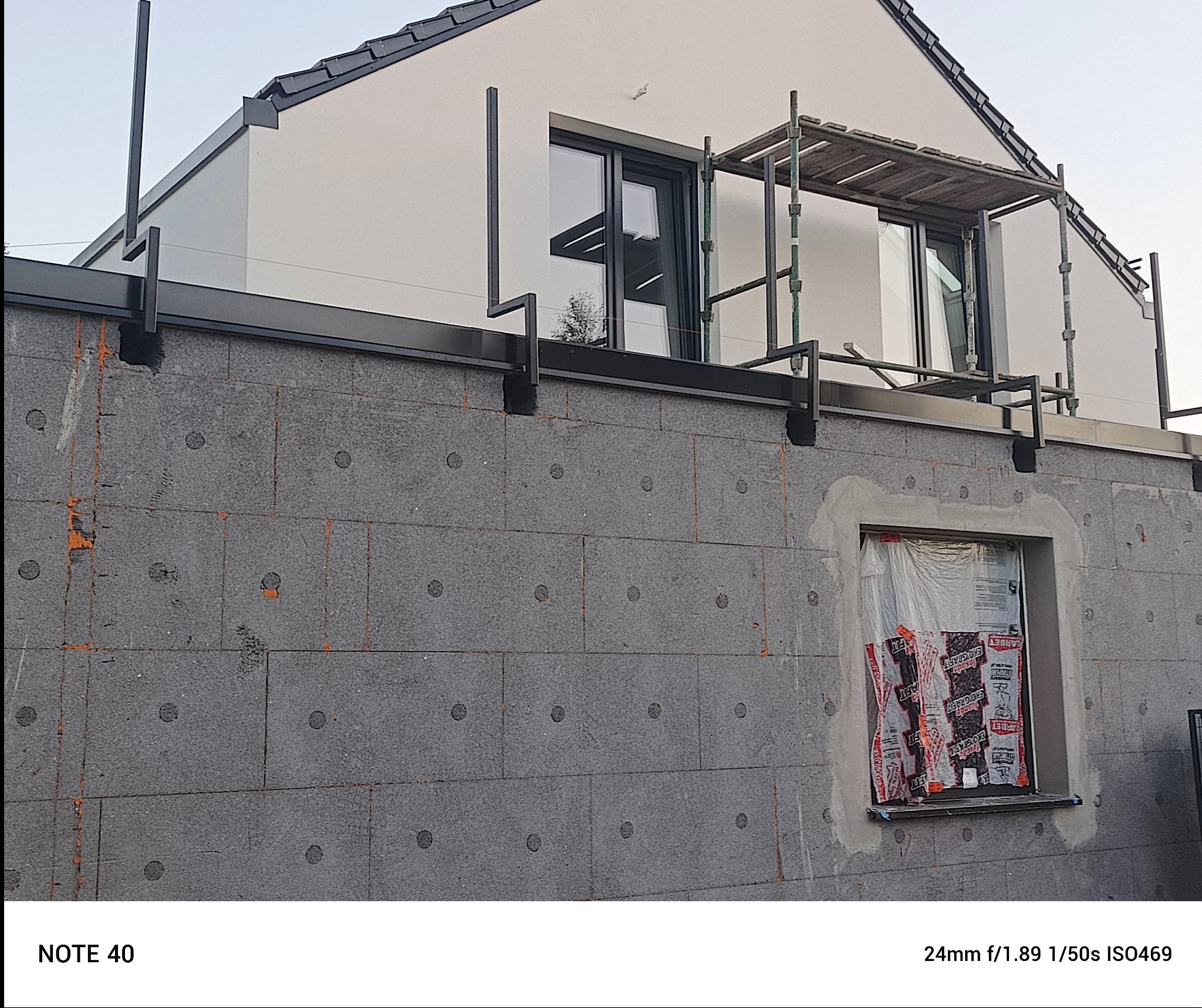 Szara elewacja w trakcie ocieplania, widoczne płyty izolacyjne z charakterystycznymi wgłębieniami, okno zabezpieczone folią, fragment rusztowania i konstrukcji dachu.