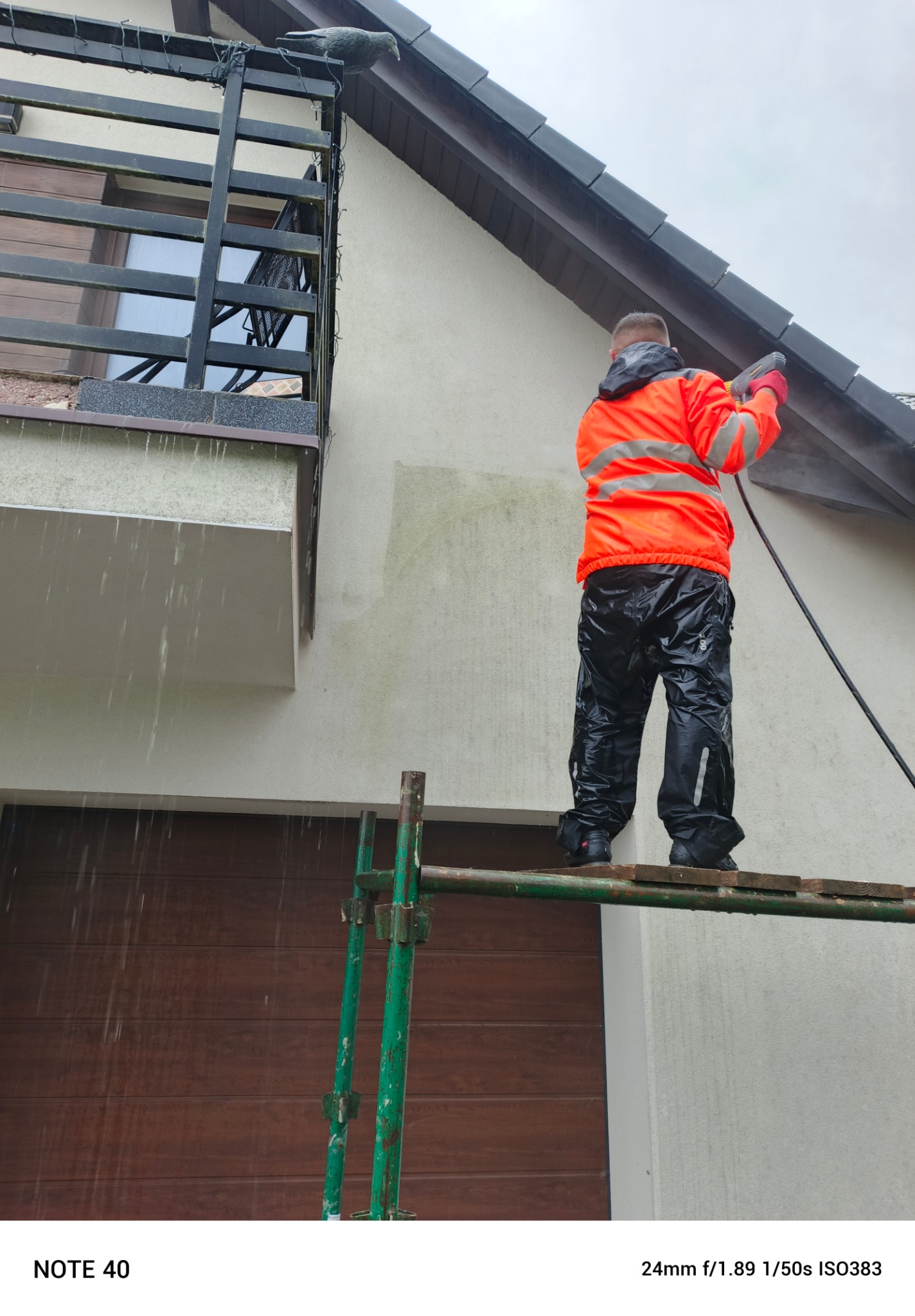 Pracownik w pomarańczowej kurtce czyści elewację domu za pomocą myjki ciśnieniowej, stojąc na rusztowaniu. Widoczne zabrudzenia na ścianie przed czyszczeniem.