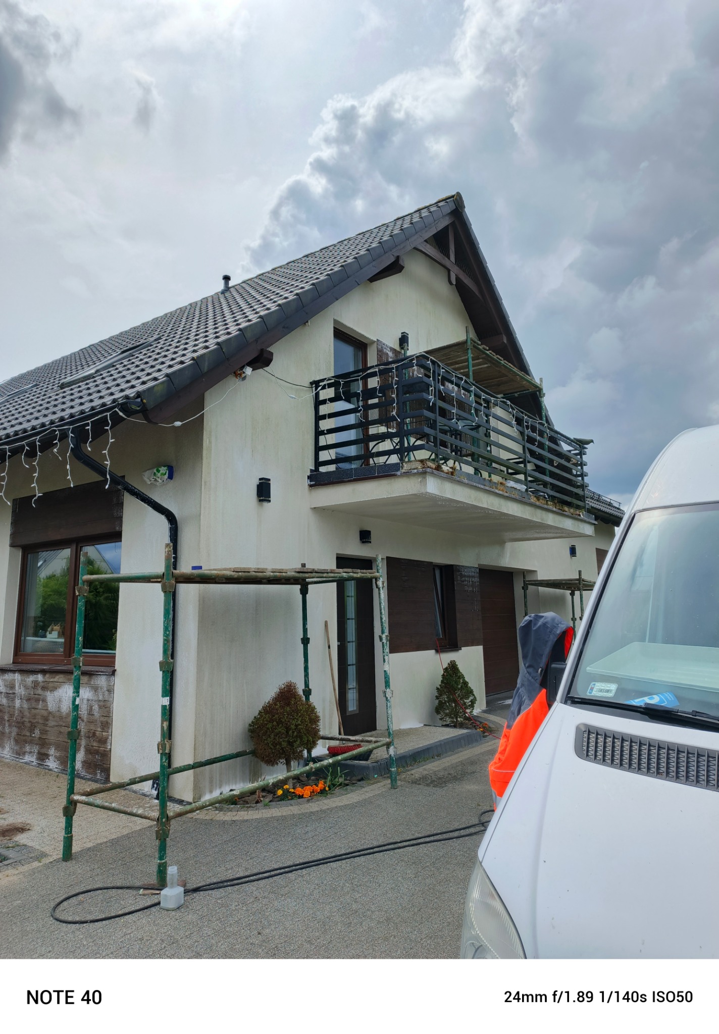 Dom z rusztowaniem przed elewacją, widoczny fragment samochodu. Ślady zabrudzeń na ścianie sugerują przygotowanie do mycia lub malowania.