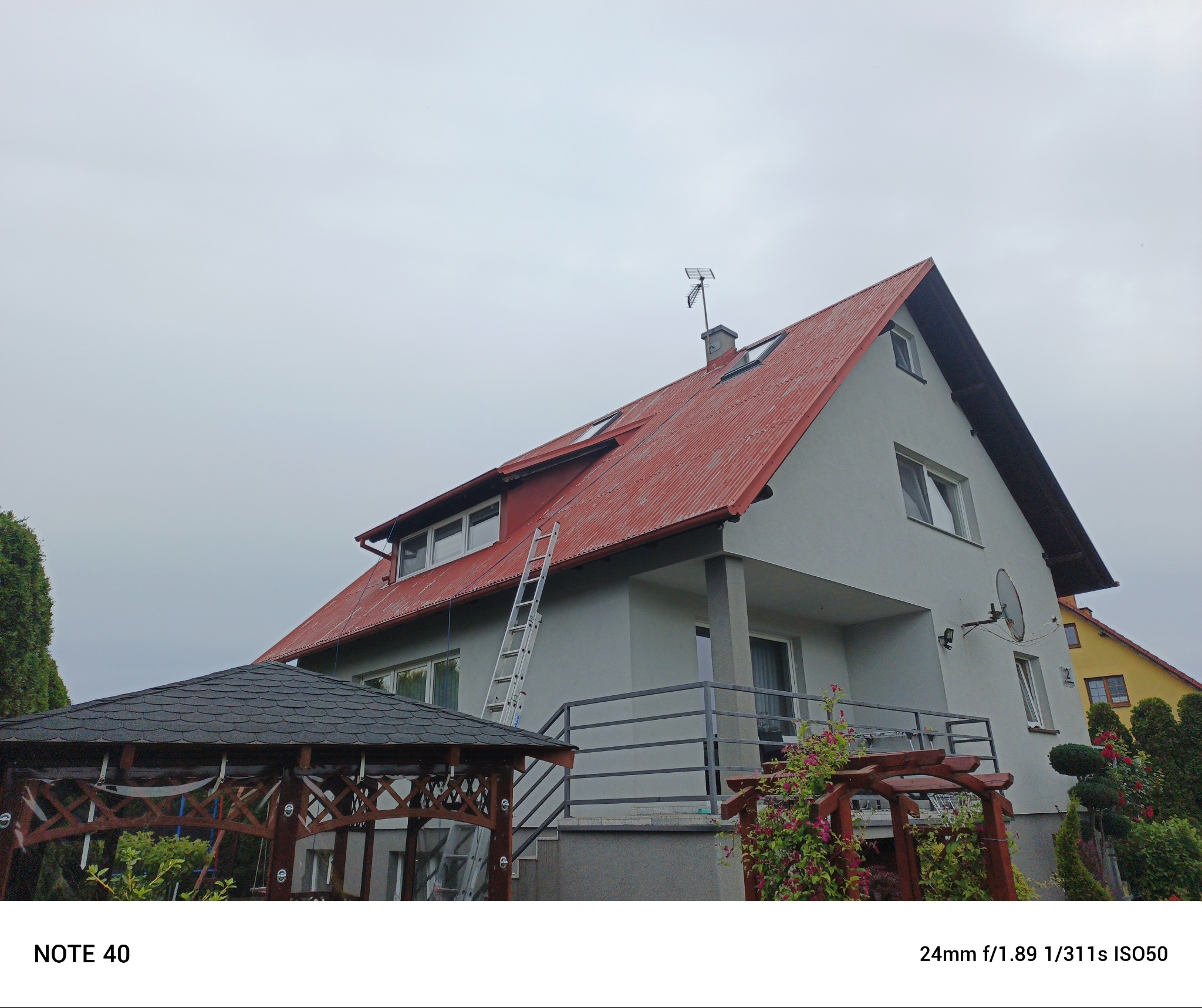 Dom z czerwonym dachem i drabiną opartą o ścianę. Widoczny fragment altany ogrodowej. Pochmurne niebo w tle.