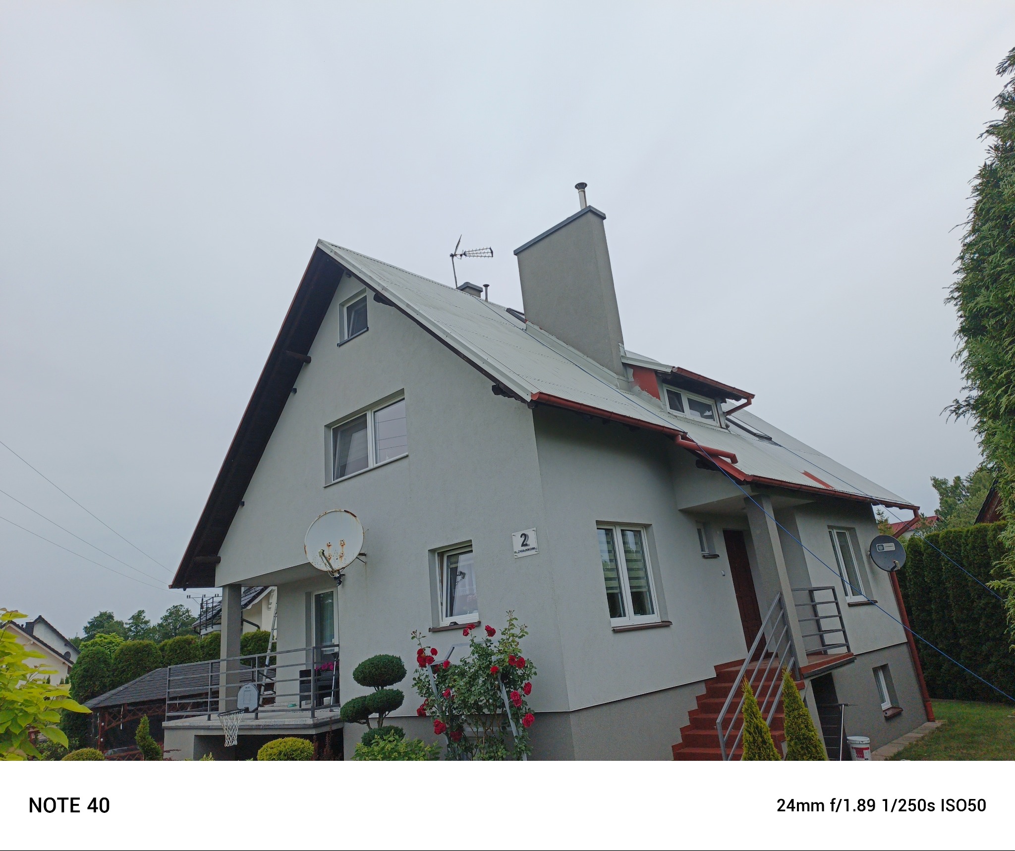 Dom jednorodzinny z szarą elewacją i metalowym dachem, widoczny z poziomu gruntu. Na dachu antena, a na elewacji numer domu i satelita. Przed domem rosną krzewy róż.