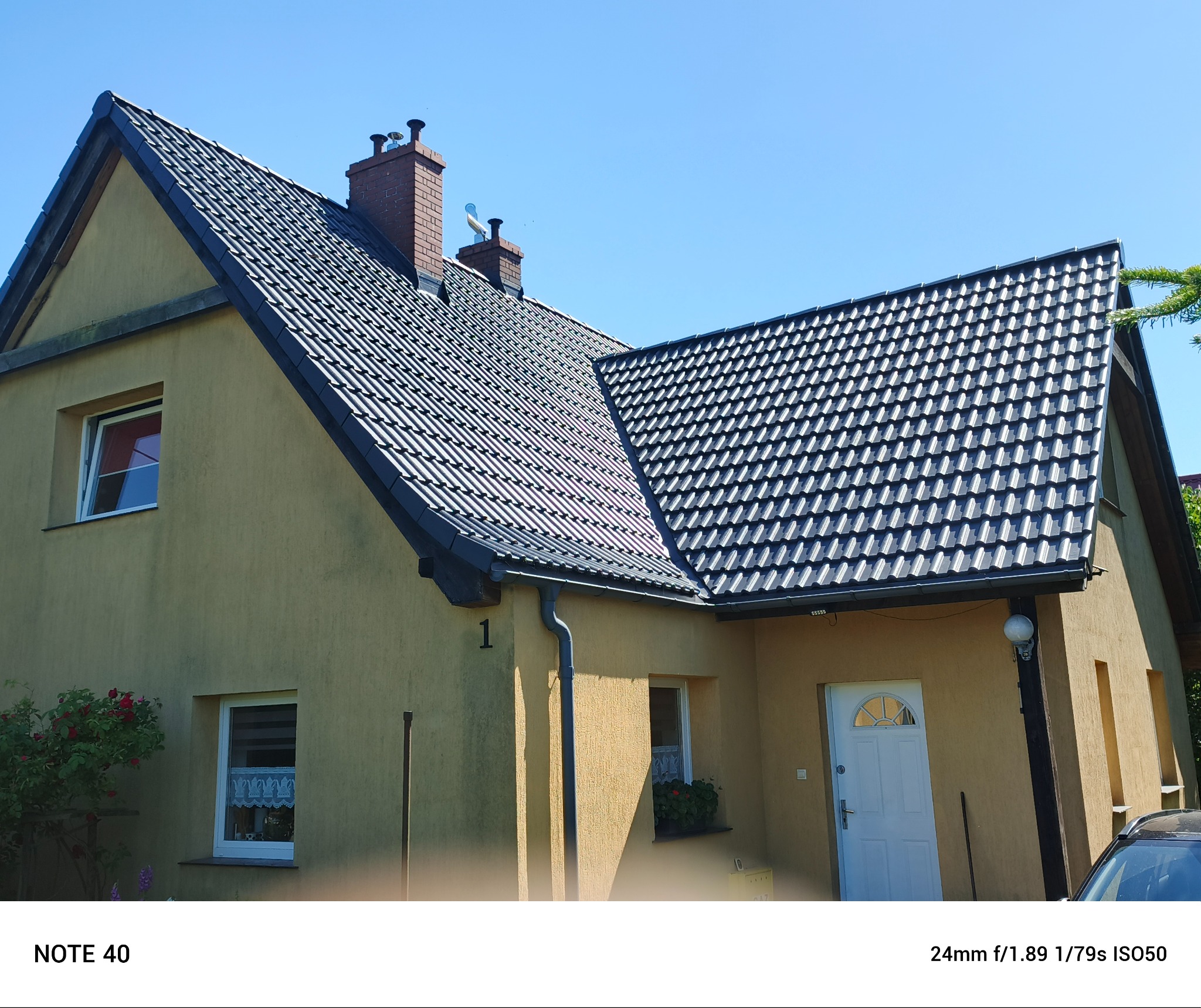 Dom jednorodzinny z ciemną dachówką w słoneczny dzień. Widoczne kominy, okna z firankami i elewacja w odcieniach beżu. Ujęcie z dołu, podkreślające geometrię dachu.