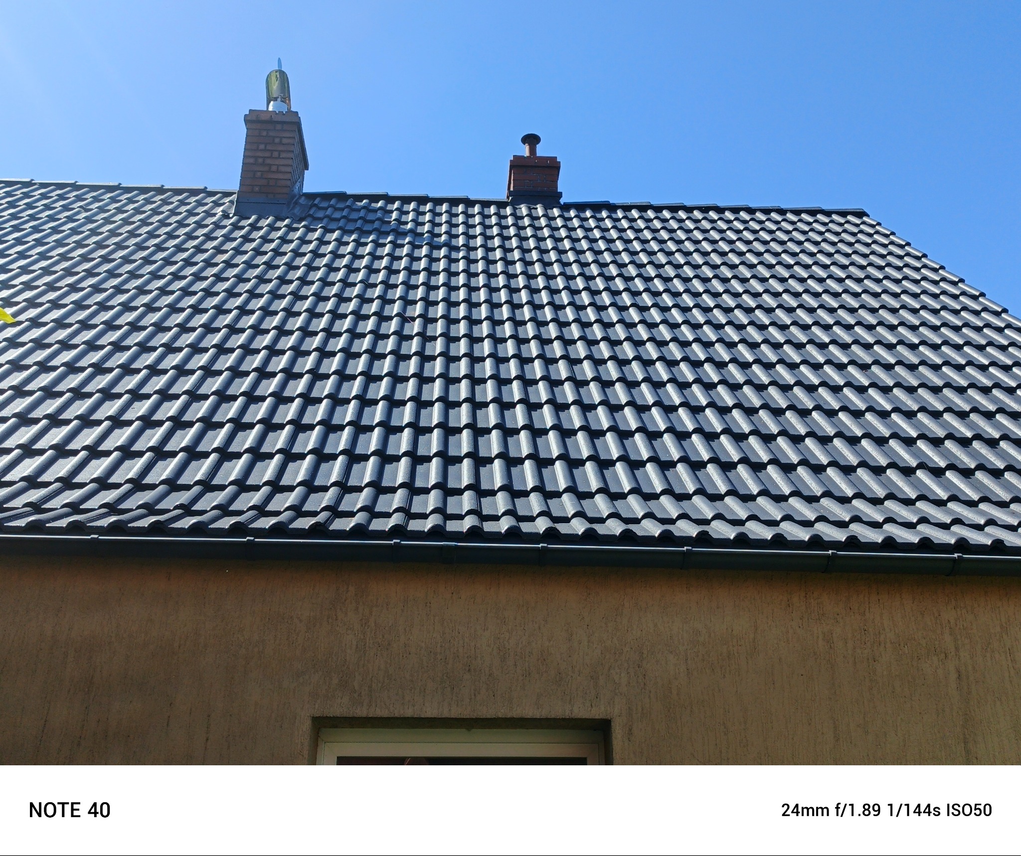 Ciemnoszara dachówka ceramiczna na skośnym dachu domu jednorodzinnego, widoczne dwa kominy z elementami wentylacyjnymi, błękitne niebo w tle.