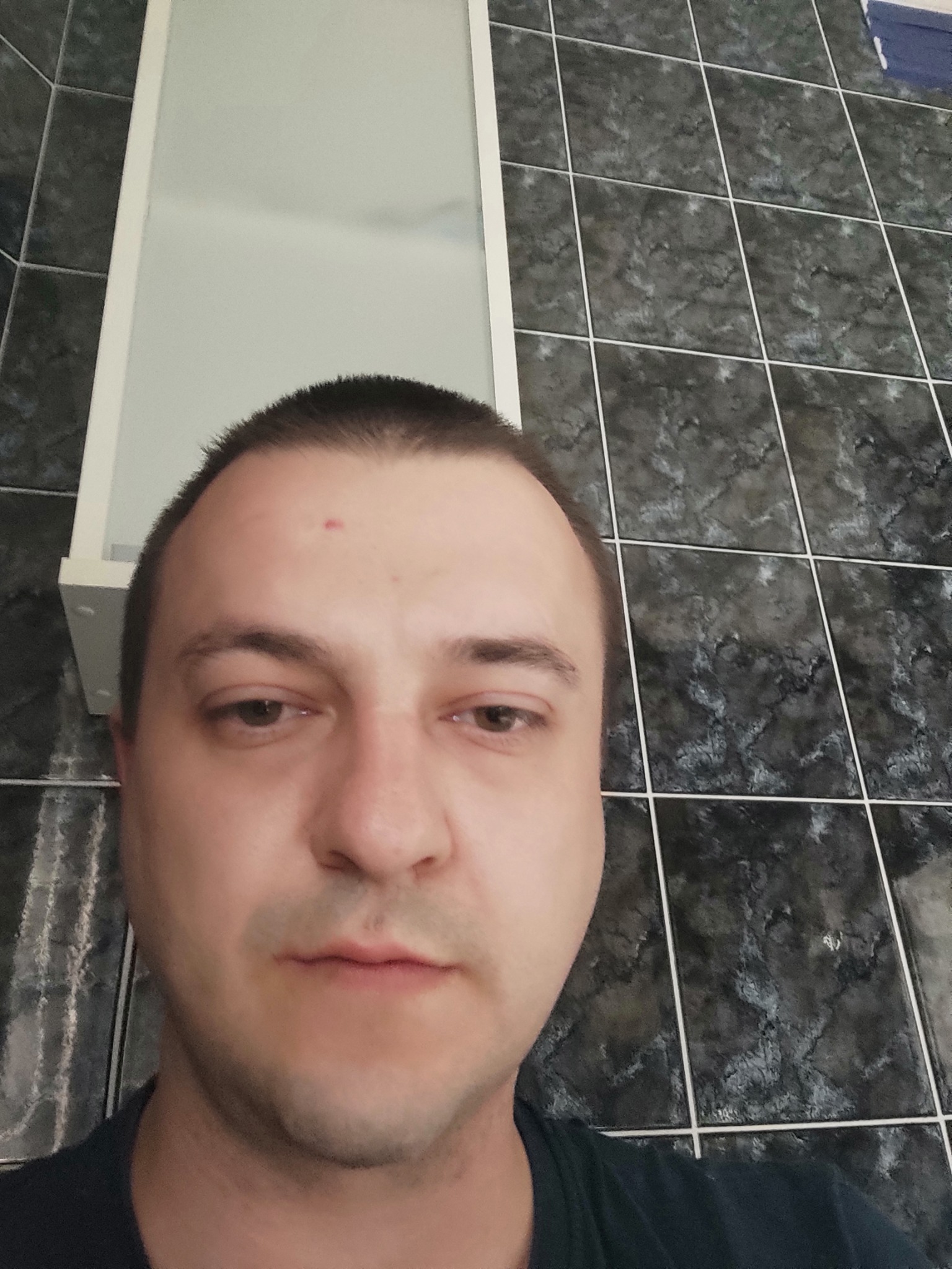 Autoportret mężczyzny na tle ściany z ciemnych płytek w łazience, widoczny fragment szafki z lustrem.