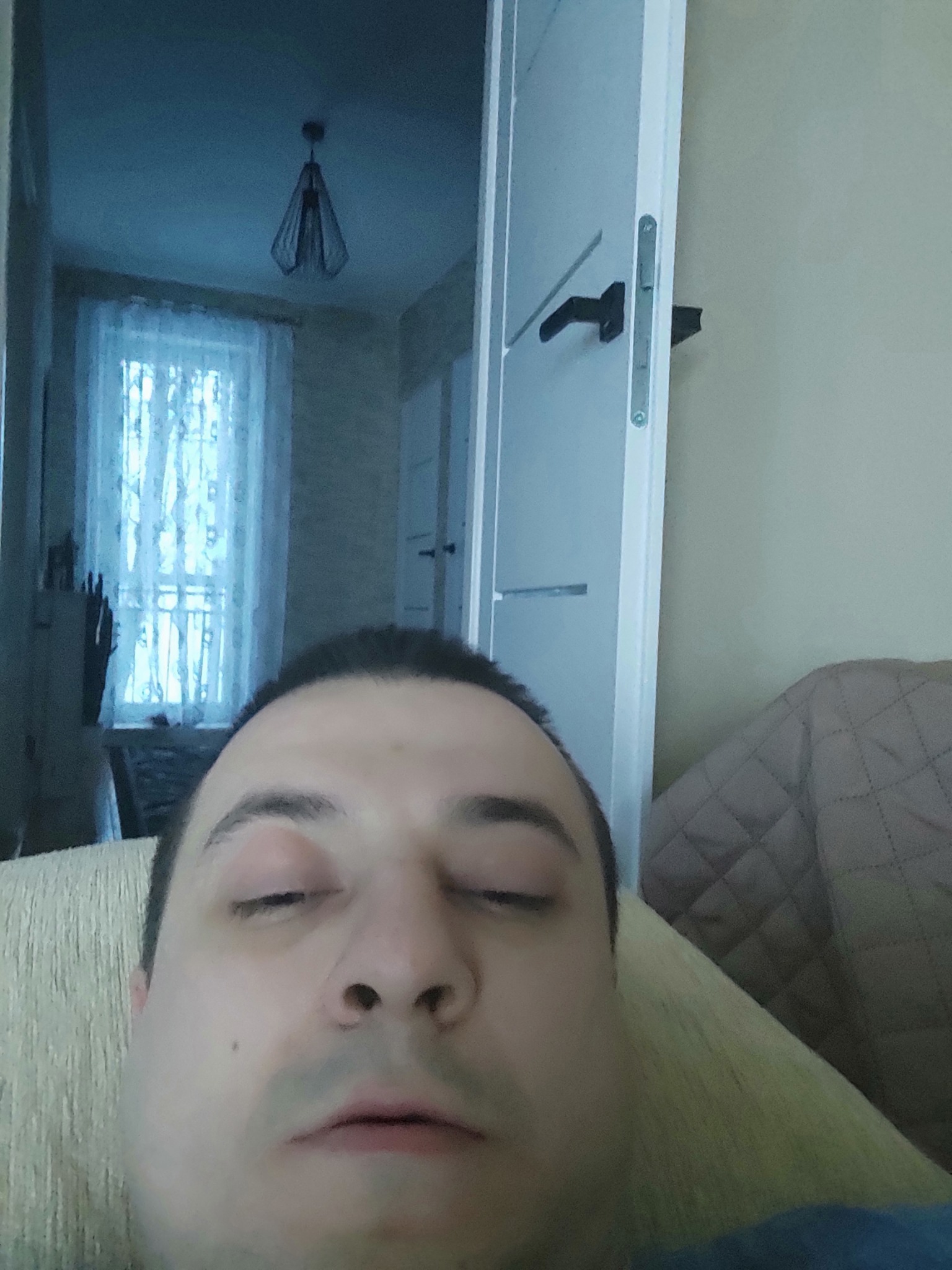 Mężczyzna z zamkniętymi oczami leżący na kanapie w słabo oświetlonym pokoju z widocznym oknem z firanką i lampą wiszącą.