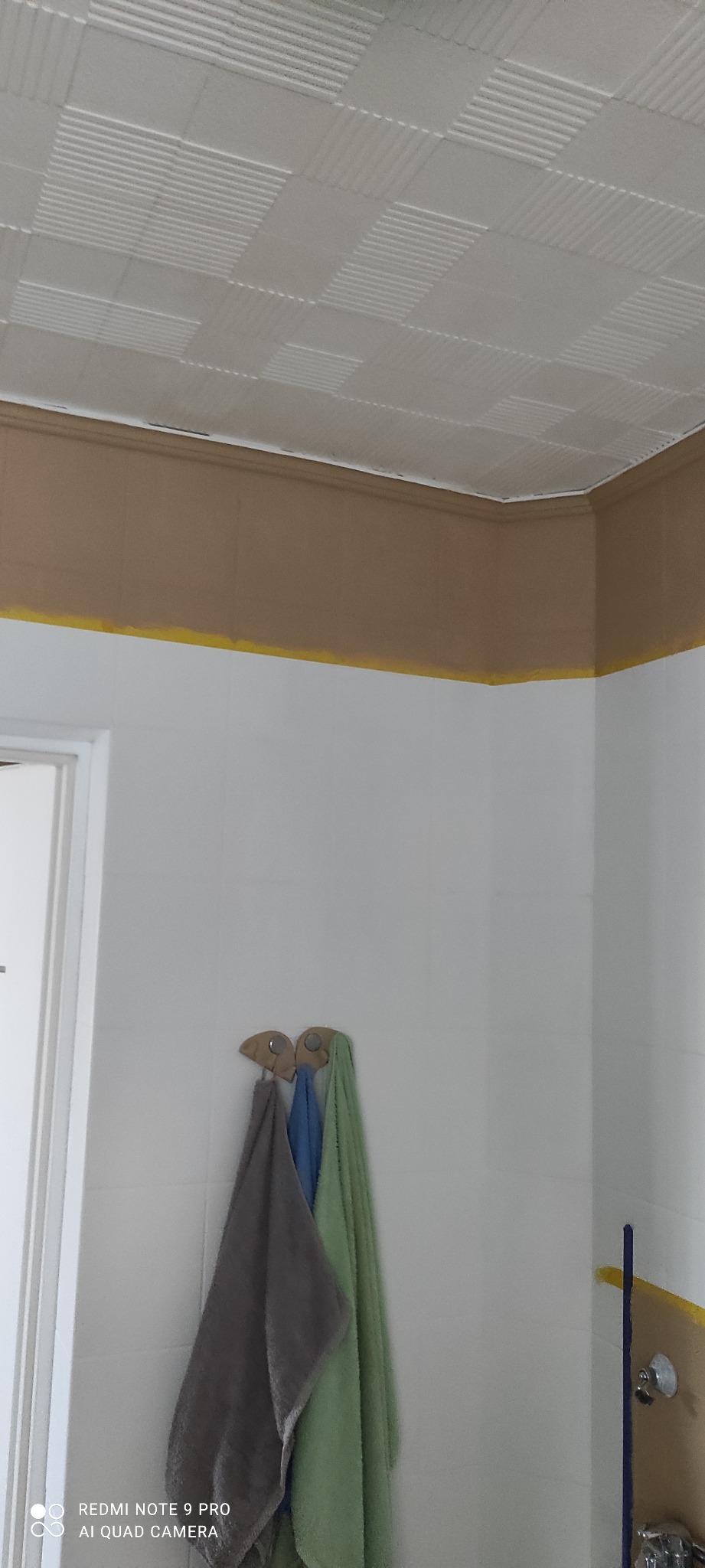 Fragment odnowionej łazienki z widocznym sufitem kasetonowym, pomalowaną górną partią ścian na kolor beżowy z żółtą linią odcięcia, oraz trzema ręcznikami wiszącymi na haczykach.