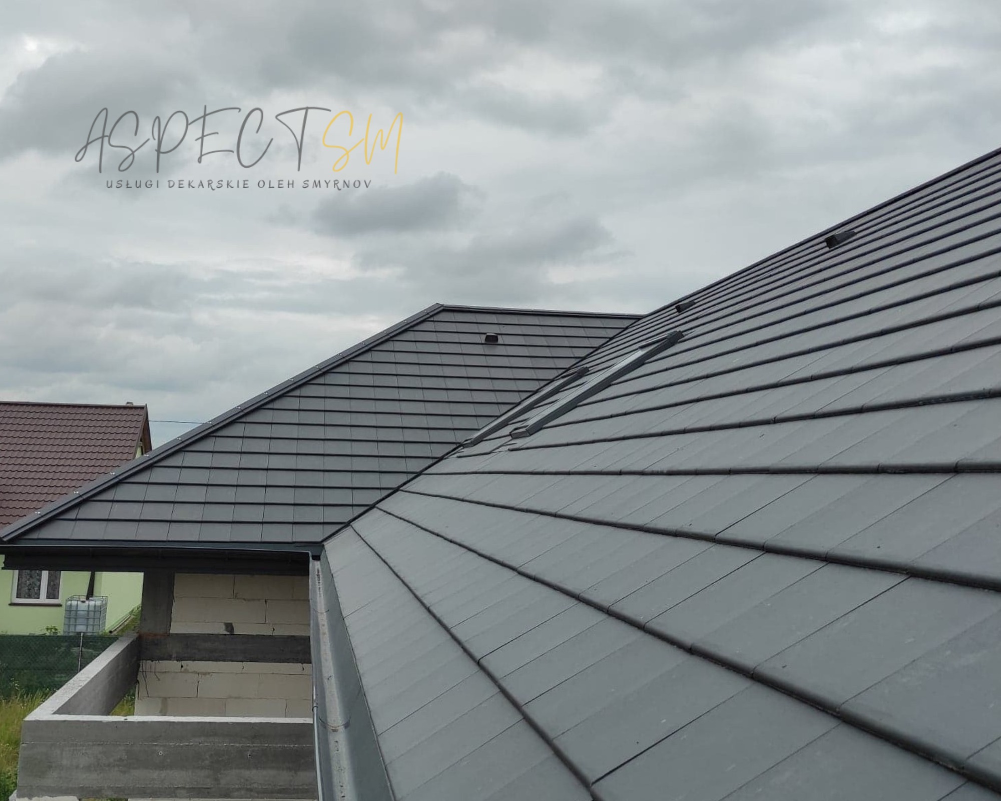 Nowoczesny, grafitowy dach z dachówki ceramicznej z oknami dachowymi, widok z góry na tle pochmurnego nieba, częściowo widoczna rynna i fragment budynku w trakcie budowy.