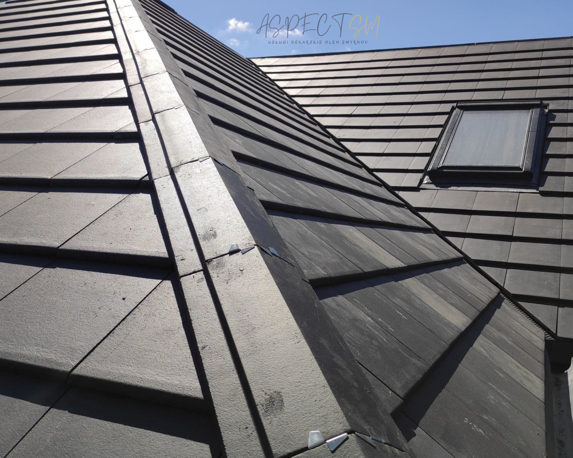 Ciemnoszary dach z dachówki ceramicznej z oknem dachowym, widoczne obróbki blacharskie i logo firmy dekarskiej w rogu zdjęcia.