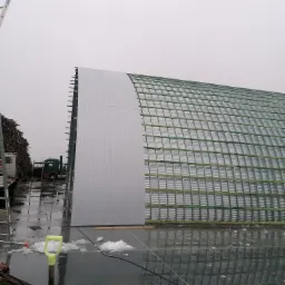 Konstrukcja łukowego dachu z metalowych paneli, częściowo pokryta, widoczna drabina oparta o bok budynku, mokra nawierzchnia z resztkami śniegu.