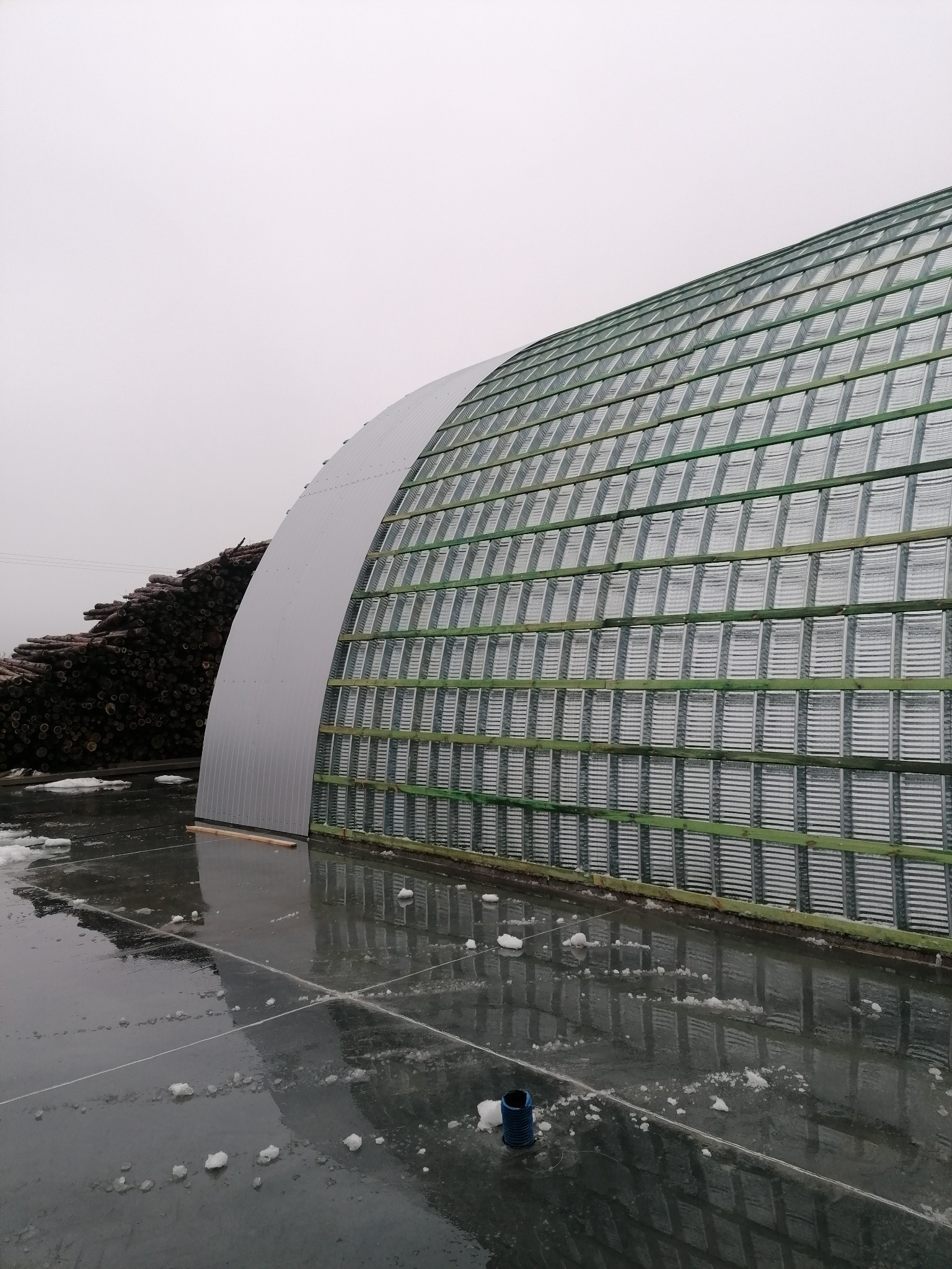 Nowoczesna, łukowa konstrukcja z metalowym poszyciem w trakcie budowy, widoczne drewniane elementy konstrukcyjne, na pierwszym planie mokra powierzchnia z resztkami śniegu, w tle stos drewna.