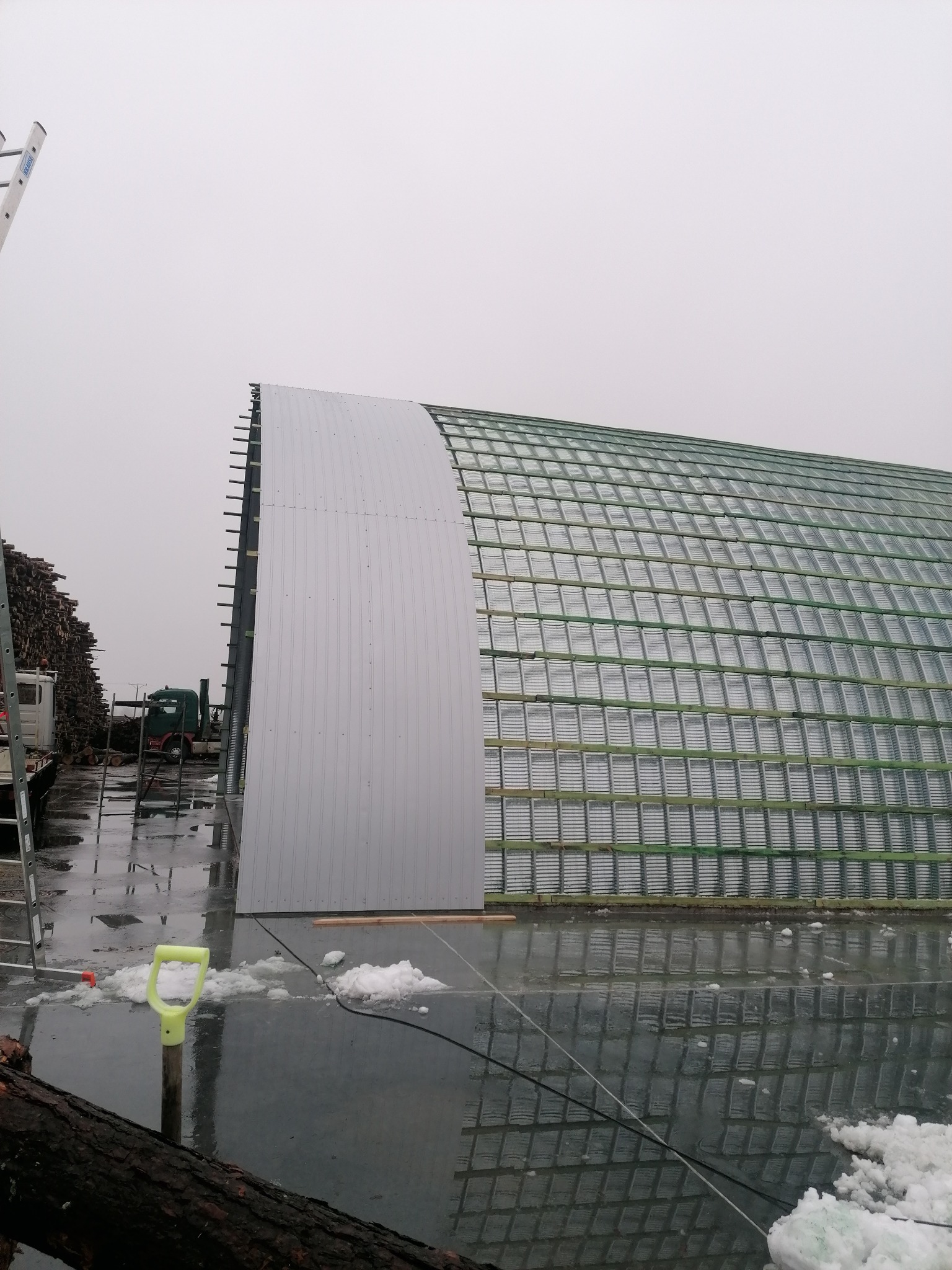 Konstrukcja łukowego dachu z metalowych paneli, częściowo pokryta, widoczna drabina oparta o bok budynku, mokra nawierzchnia z resztkami śniegu.