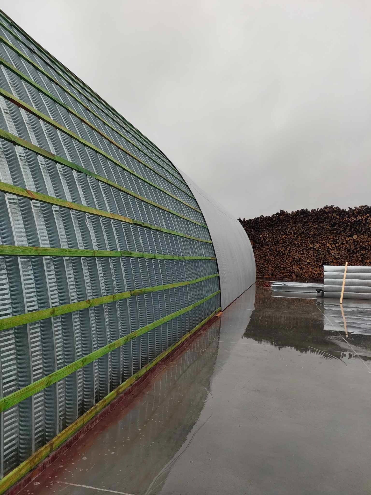 Konstrukcja łukowego dachu z blachy falistej, zielone łaty drewniane, mokra betonowa posadzka, w tle stos drewna opałowego pod pochmurnym niebem.