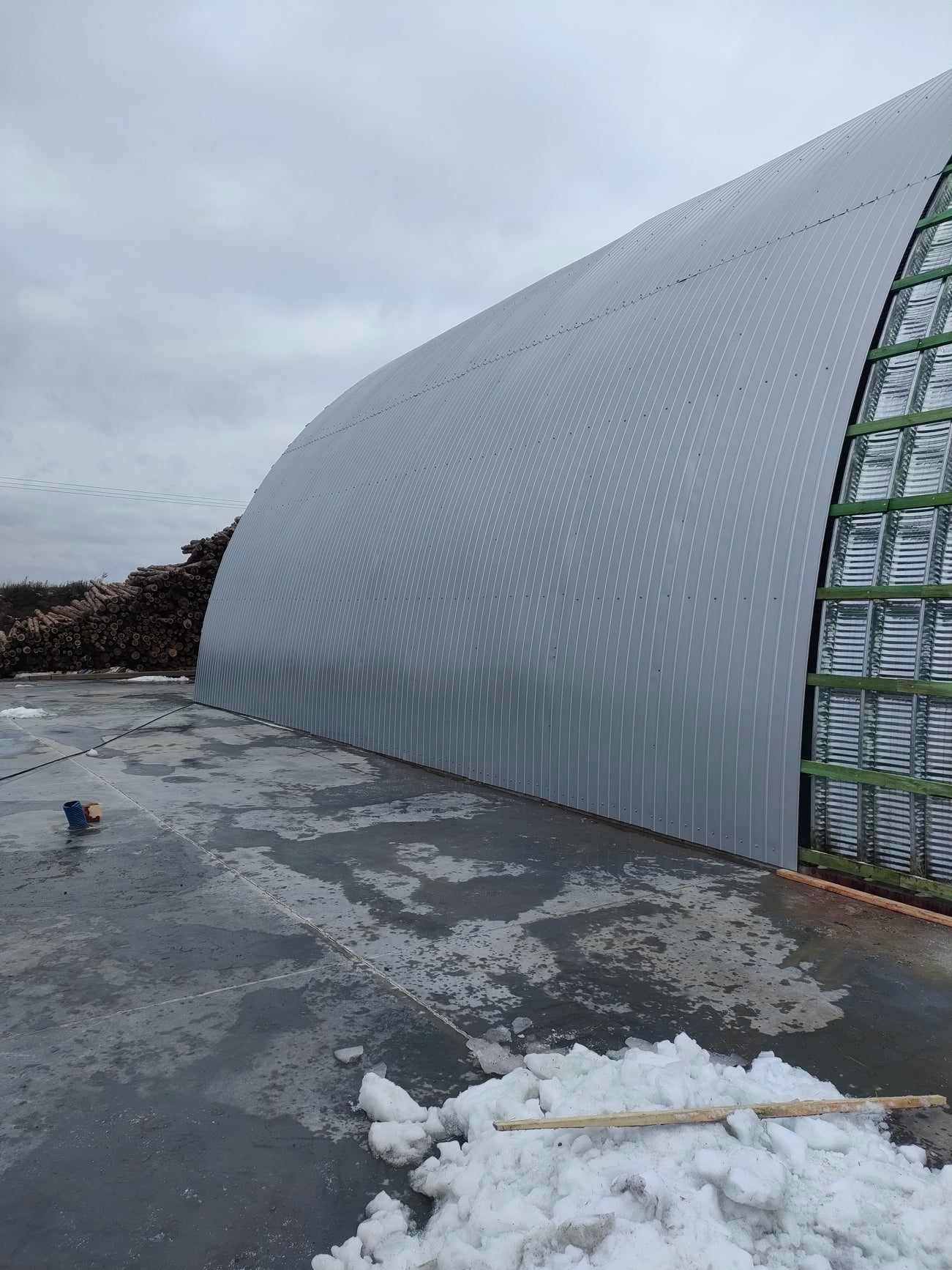 Konstrukcja łukowa z blachy falistej na betonowej płycie, widoczne drewniane elementy szkieletu i resztki śniegu.