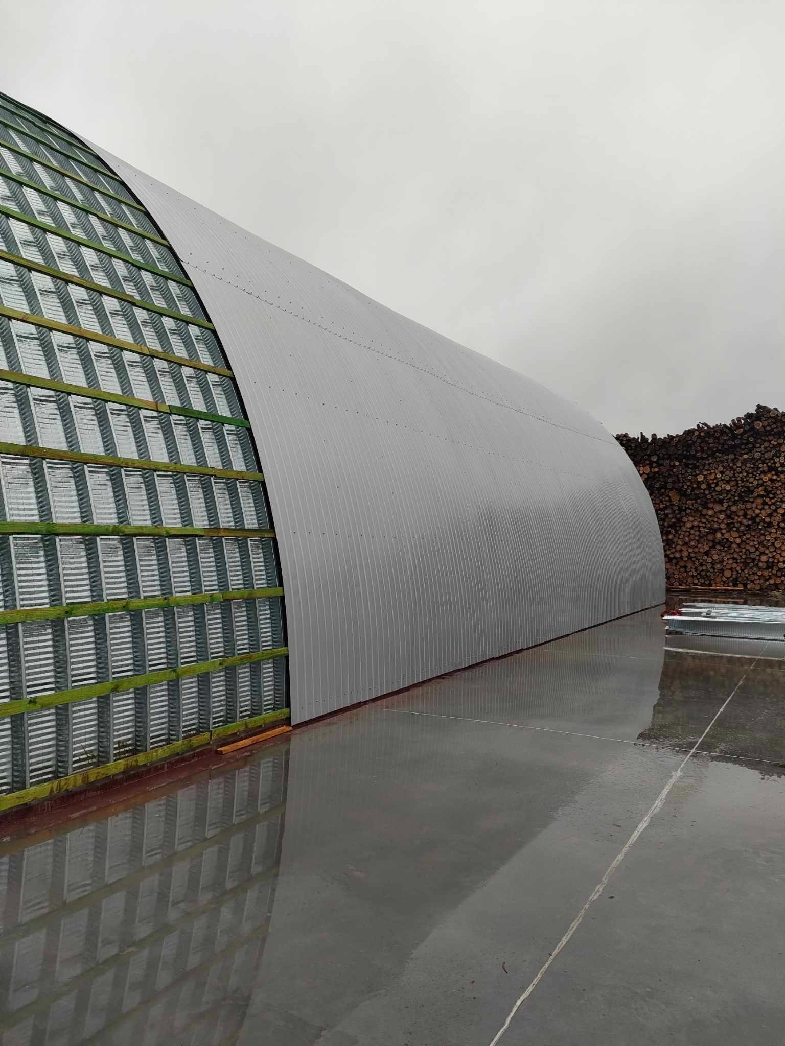 Konstrukcja łukowego dachu z blachy trapezowej w trakcie montażu, widoczne drewniane łaty, na zewnątrz stos drewna, mokra betonowa posadzka.