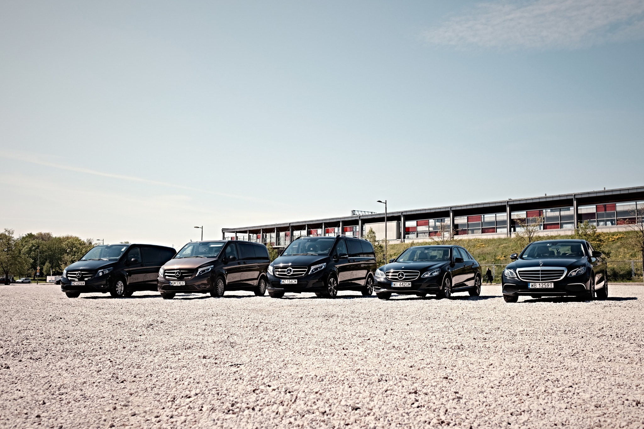 Rząd pięciu luksusowych samochodów Mercedes-Benz na żwirowym parkingu przed budynkiem z czerwonymi akcentami okiennymi, w słoneczny dzień.