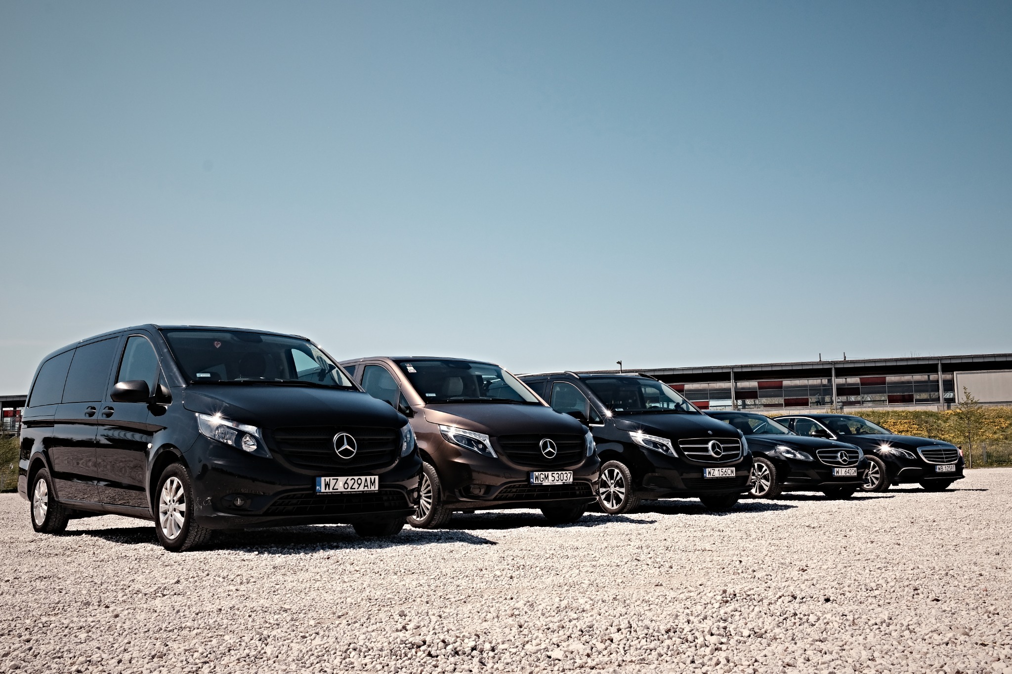 Flota pojazdów marki Mercedes-Benz w kolorach czarnym i brązowym, stojąca na żwirowym parkingu pod błękitnym niebem.