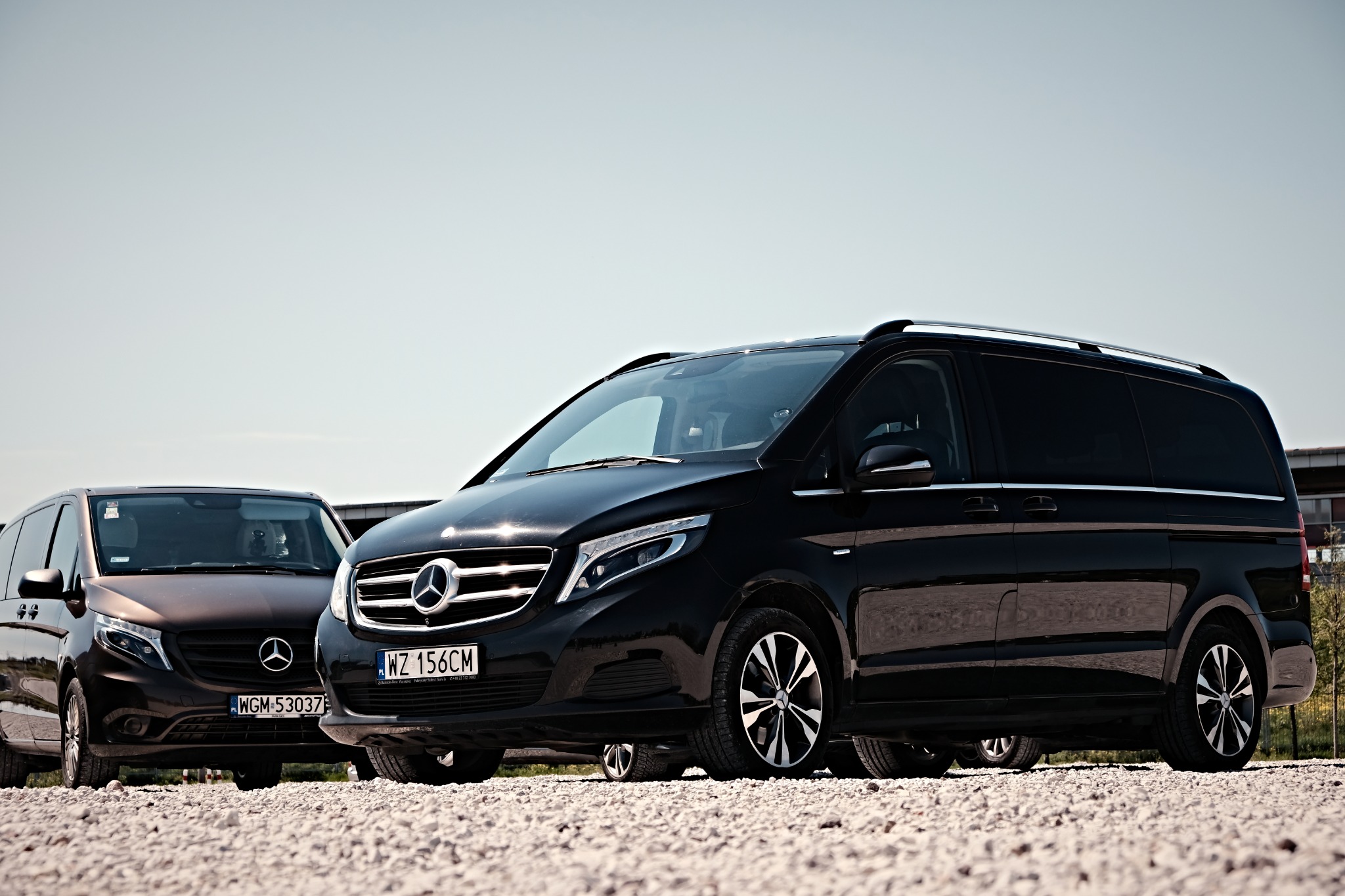 Dwa czarne busy Mercedes V-klasa zaparkowane na żwirowym parkingu, widoczne tablice rejestracyjne.