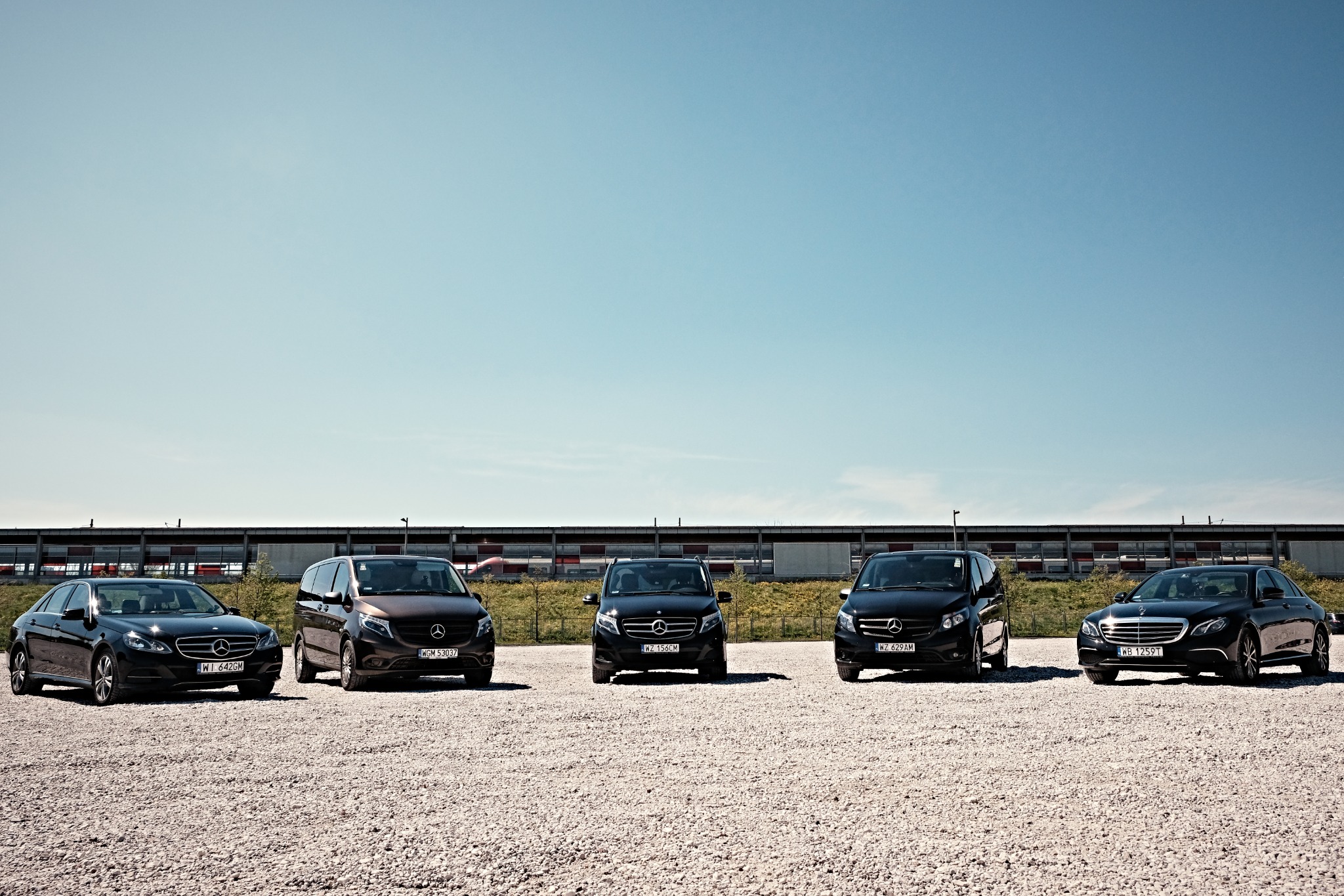 Flota pięciu samochodów Mercedes-Benz (sedany i vany) ustawiona w rzędzie na żwirowym parkingu przed budynkiem w słoneczny dzień.