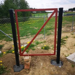 Surowa, czerwona rama bramy z ukośnym wzmocnieniem, tymczasowo podparta cegłami, osadzona na czarnych słupkach w świeżym betonie, z widocznym łączeniem z siatką ogrodzeniową w tle, na tle łąki...