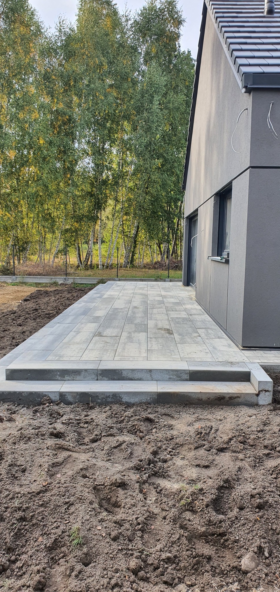 Nowoczesny taras z szarej kostki brukowej przylegający do domu z ciemną elewacją, widoczne dwa stopnie wejściowe i przygotowany teren pod trawnik.