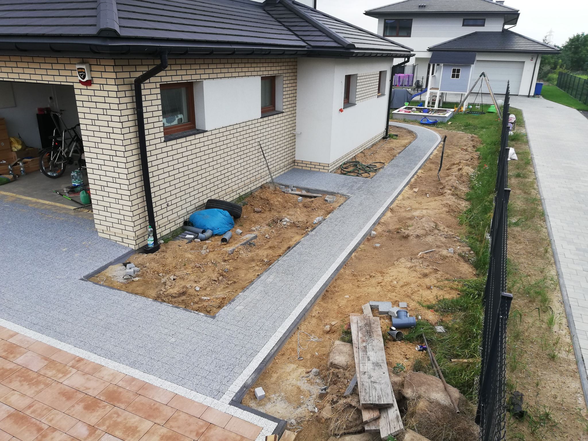 Fragment posesji w trakcie budowy z częściowo ułożoną szarą kostką brukową na podjeździe i chodniku, widoczne przygotowane podłoże, narzędzia i materiały budowlane, dom w tle.