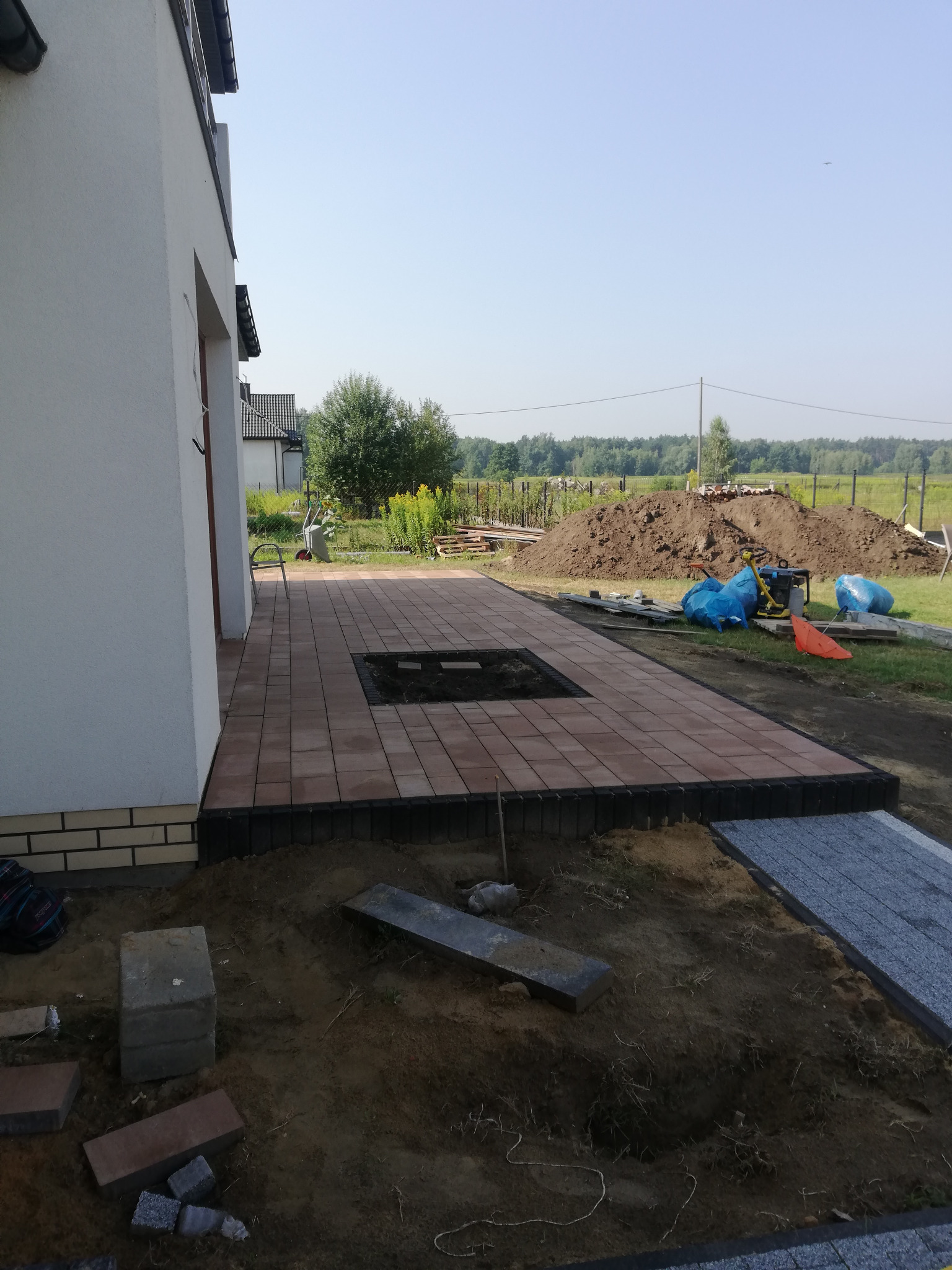 Częściowo ułożony taras z brązowej kostki brukowej przy białym domu, widoczny fragment przygotowywanego podłoża i narzędzia budowlane.