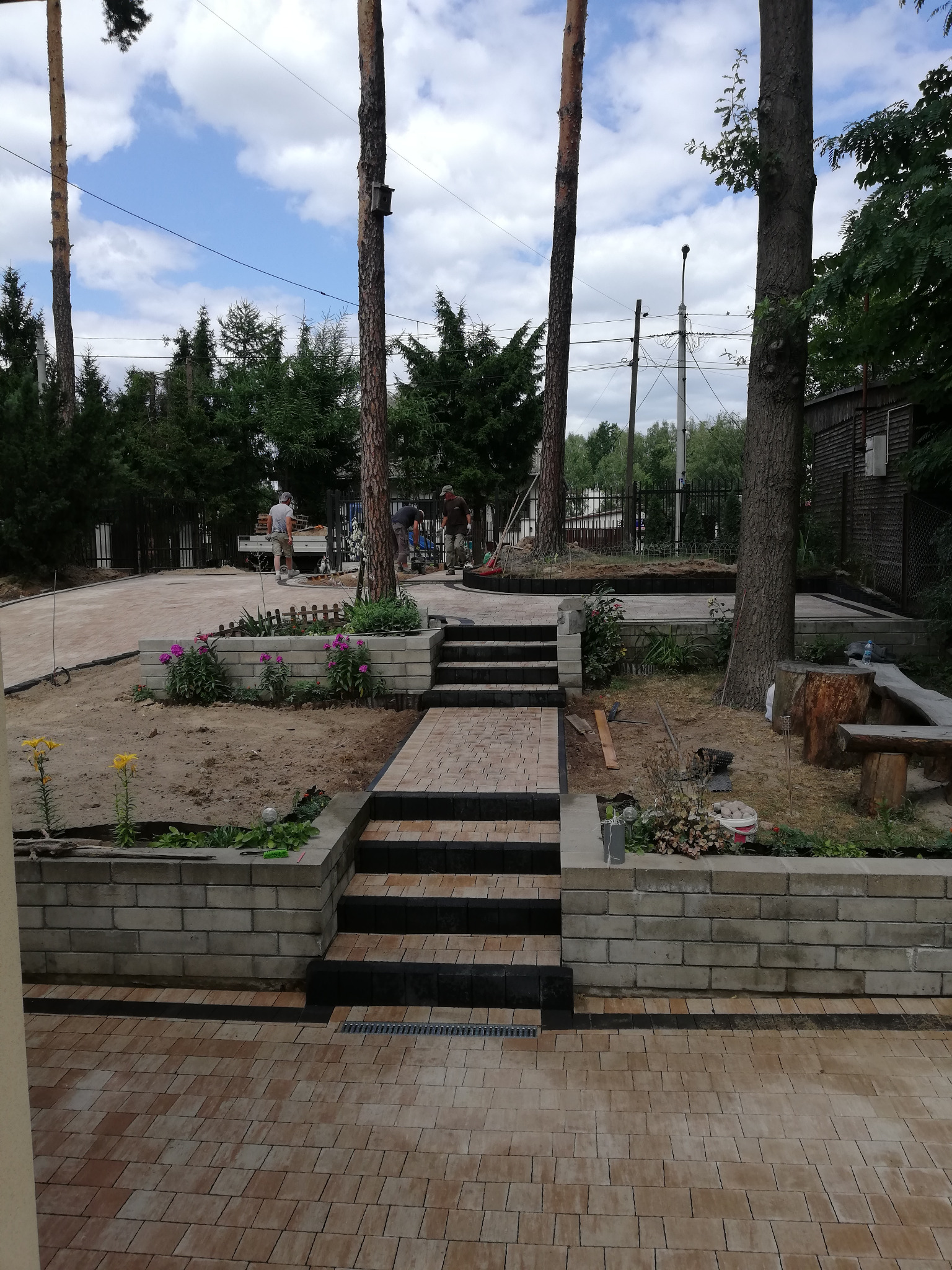 Ułożona kostka brukowa w odcieniach beżu i brązu prowadząca do schodów z czarnymi krawędziami, z pracownikami w tle i elementami zieleni w ogrodzie.