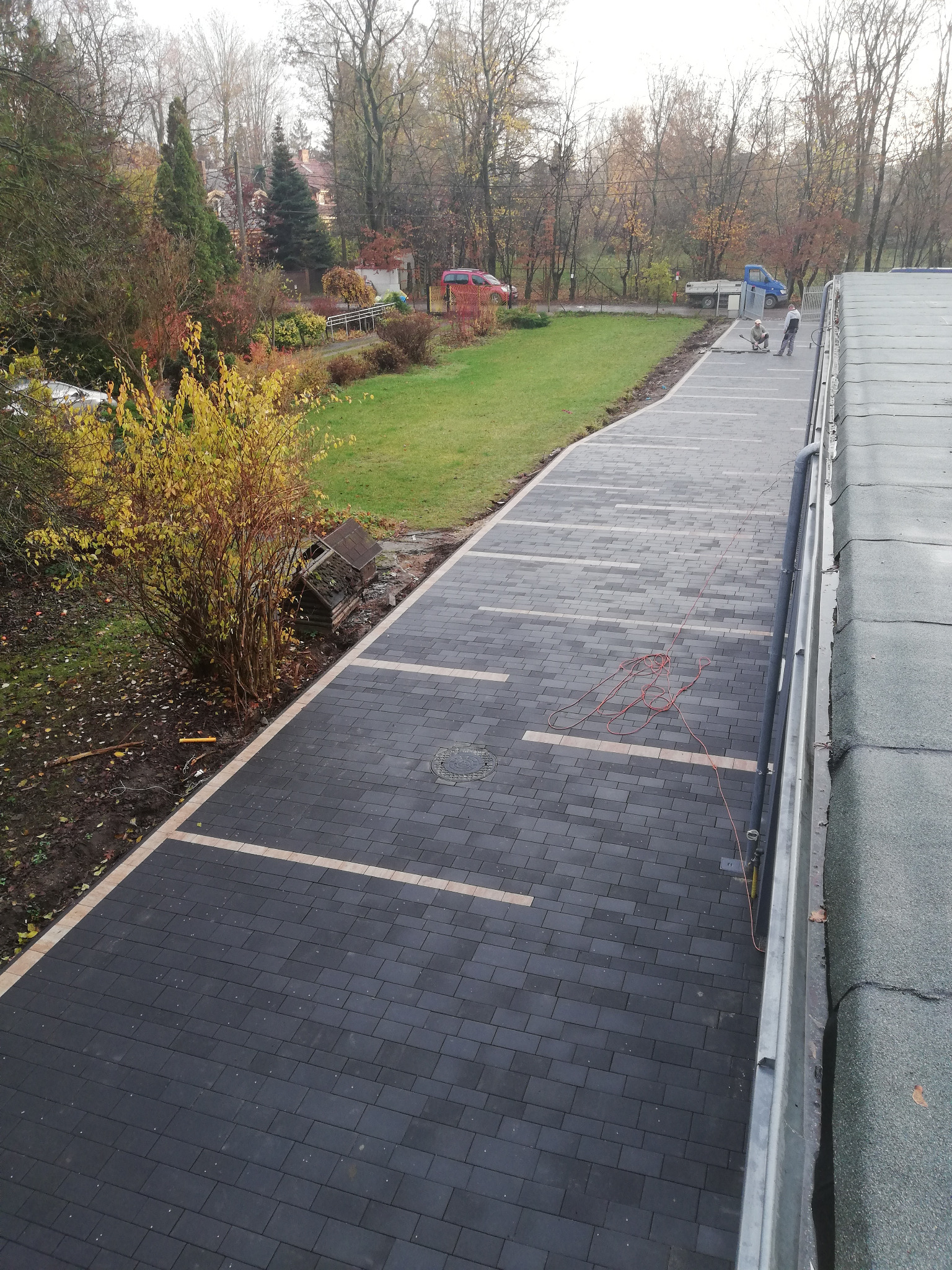 Układanie ciemnoszarej kostki brukowej z jasnymi pasami w Piasecznie, widok z góry, dwóch pracowników w oddali, na obrzeżach widoczne drzewa i krzewy.