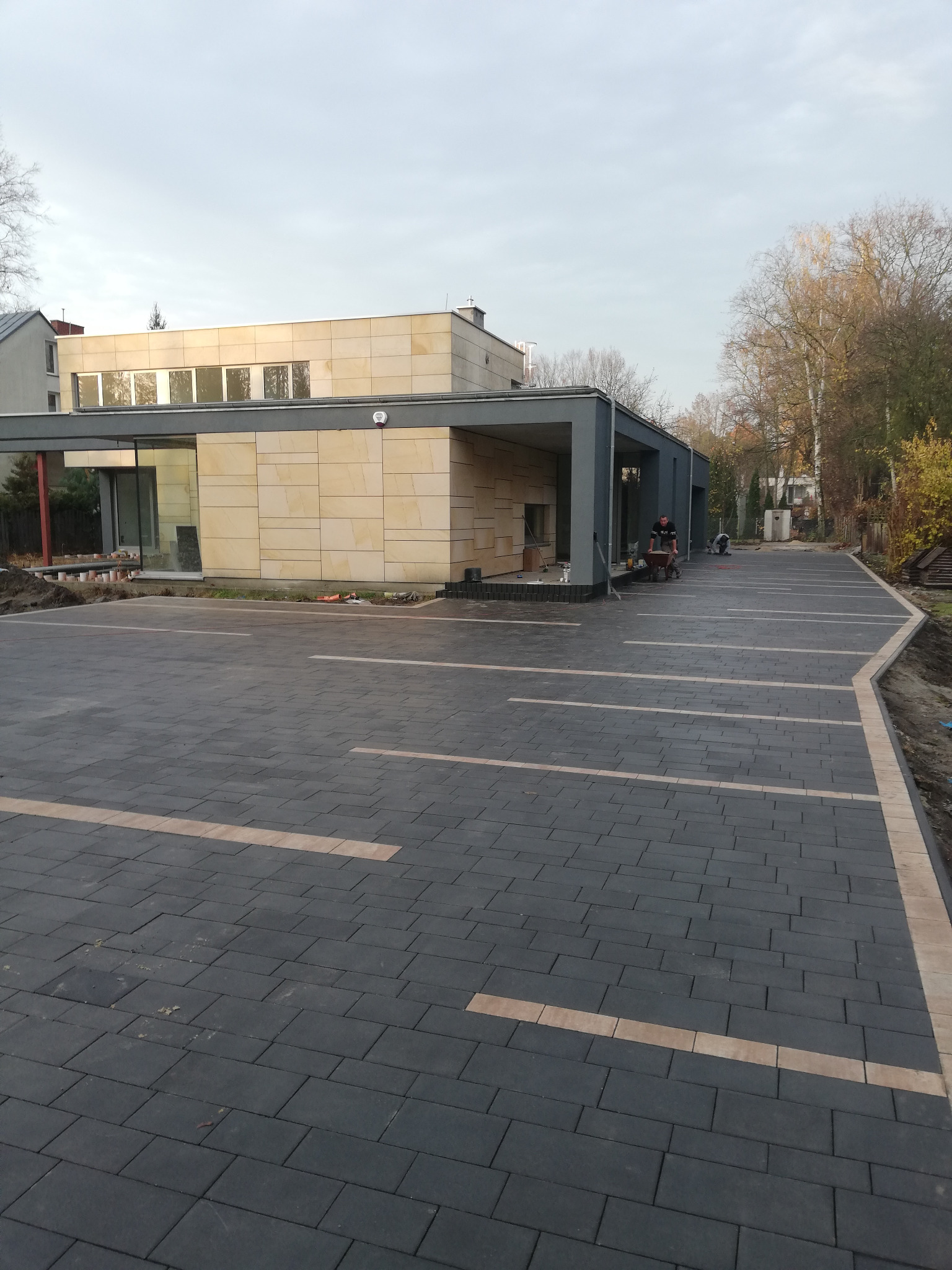 Nowoczesna posesja z ciemną kostką brukową z jasnymi wstawkami, widoczni pracownicy z taczką i narzędziami w tle, budynek z jasnej cegły w tle.