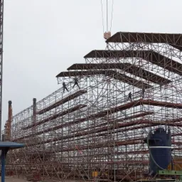 Montaż wielopoziomowego rusztowania z metalowych rur na placu budowy, widoczni robotnicy w oddali i dźwig w tle.