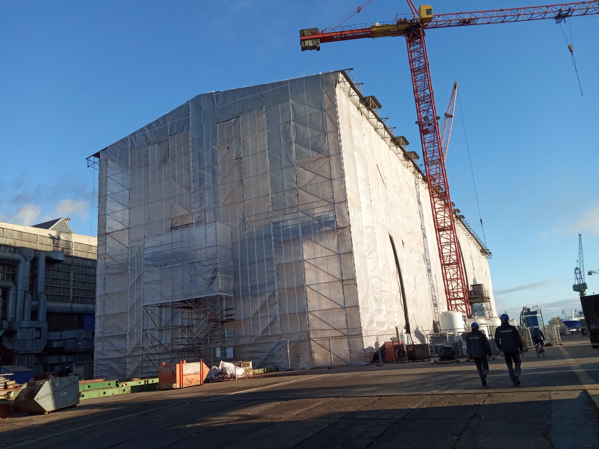 Duży budynek w trakcie budowy, osłonięty białą plandeką i rusztowaniami, z dwoma robotnikami w oddali i żurawiem budowlanym na tle błękitnego nieba.