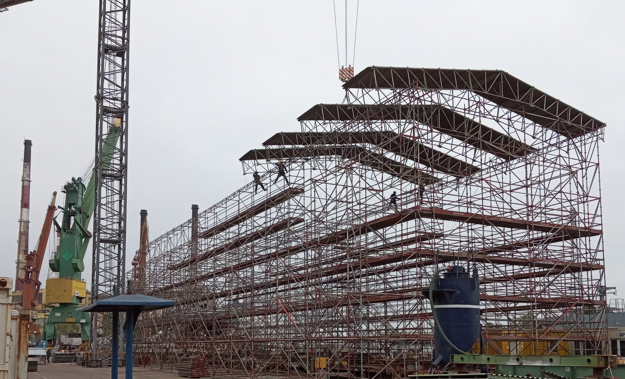 Montaż wielopoziomowego rusztowania z metalowych rur na placu budowy, widoczni robotnicy w oddali i dźwig w tle.