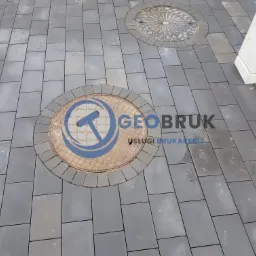 Ułożona szara kostka brukowa z dwoma studzienkami kanalizacyjnymi, jedna z logo firmy brukarskiej 'GEOBRUK', druga z dekoracyjnym wzorem.