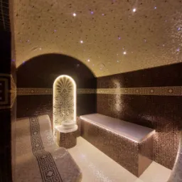 Mozaikowa łazienka z podświetlanym sufitem imitującym gwiazdy, dekoracyjnym panelem ściennym i podgrzewanymi leżankami w stylu greckim.