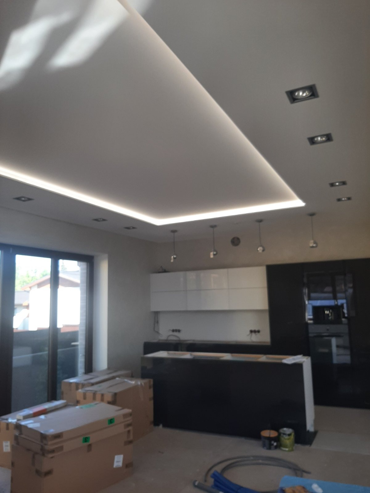 Nowoczesna kuchnia w trakcie montażu, z podwieszanym sufitem z oświetleniem LED, czarnymi i białymi szafkami, oraz zapakowanymi elementami wyposażenia na podłodze.