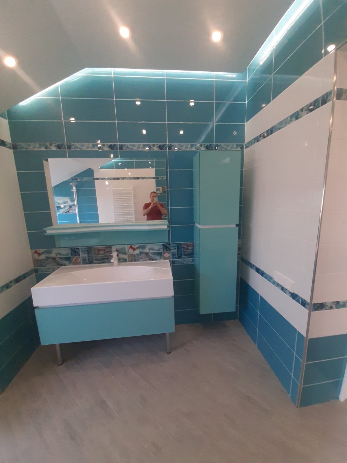 Turkusowo-biała łazienka z umywalką na szafce, lustrem, szafką wiszącą i mozaikowymi akcentami, widok z perspektywy wejścia.