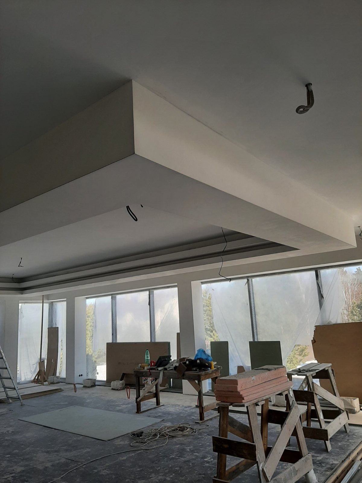 Wnętrze w trakcie remontu: widoczne płyty karton-gips na podłodze, rusztowania, okna zakryte folią ochronną i fragmenty nowo wykonanej zabudowy sufitowej z elementami instalacji elektrycznej.