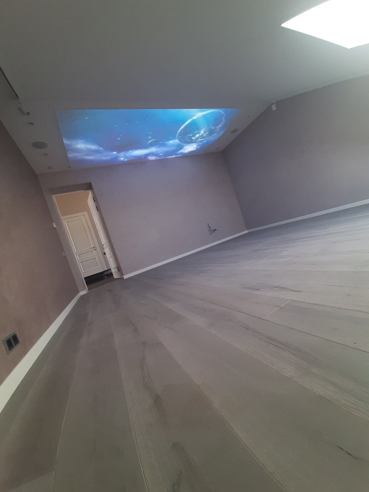 Pusty pokój z szarymi panelami podłogowymi i projekcją galaktyki na suficie, widoczne gładkie ściany w odcieniach szarości i białe listwy przypodłogowe, drzwi w tle.