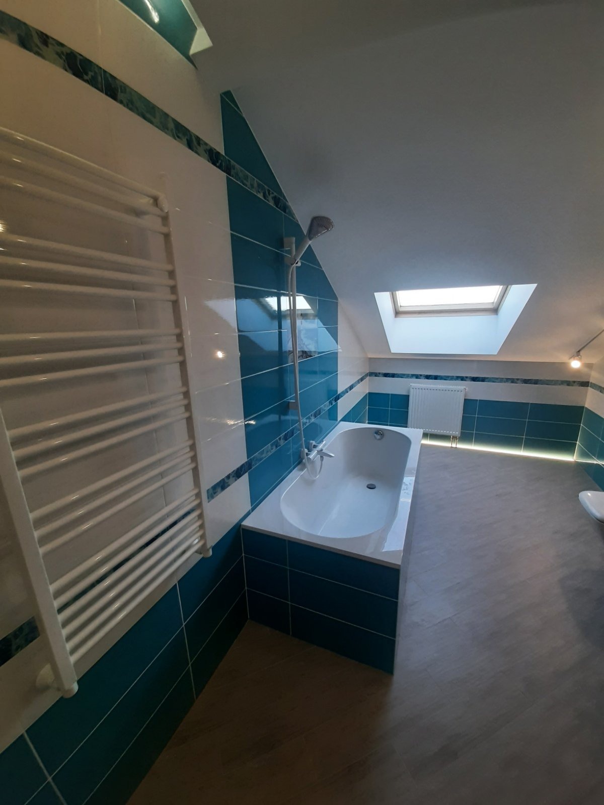 Wykończona łazienka na poddaszu z biało-turkusowymi płytkami, białą wanną, grzejnikiem drabinkowym i oknem dachowym.