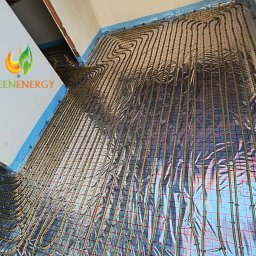 GREEN ENERGY Sp. z.o.o. Pompy Ciepła Nowicki Technika Grzewcza - Instalacja ogrzewania podłogowego z rurkami układanymi w pętle na folii termoizolacyjnej i siatce montażowej, widoczne logo firmy Green Energy na ścianie.
