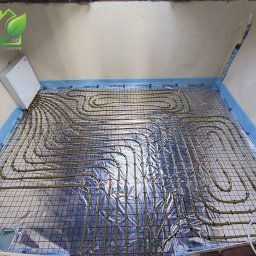 GREEN ENERGY Sp. z.o.o. Pompy Ciepła Nowicki Technika Grzewcza - Instalacja ogrzewania podłogowego: rury grzewcze ułożone na siatce z folią odblaskową, widoczna puszka elektryczna na ścianie, przygotowanie do wylewki.