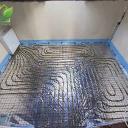 Instalacja ogrzewania podłogowego: rury grzewcze ułożone na siatce z folią odblaskową, widoczna puszka elektryczna na ścianie, przygotowanie do wylewki.