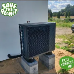 Szara pompa ciepła Panasonic, zamontowana na betonowych blokach przy białej ścianie budynku, z naklejkami 'Save the planet' i 'Eco friendly'.