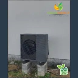 Szara pompa ciepła zamontowana na betonowych blokach przy jasnej ścianie budynku. Widoczne logo firmy Green Energy i hasło 'Bądź z nami EKO'.